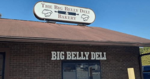 Sign for "The Big Belly Deli & Bakery" on a brick building, featuring a large window and a sloped roof.