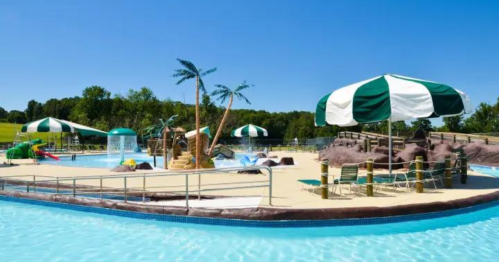 A sunny pool area featuring palm trees, umbrellas, and water slides, surrounded by a sandy beach-like setting.