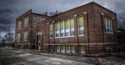 Abandoned brick school building with yellowed windows, surrounded by a gloomy sky and overgrown vegetation.