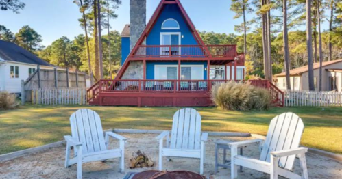 A colorful A-frame house surrounded by trees, with a fire pit and seating area in the foreground.