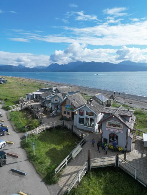 A coastal scene featuring colorful shops along a boardwalk, with a beach and mountains in the background under a blue sky.