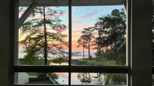 Sunset view through a window, featuring trees, a calm lake, and a pier against a colorful sky.