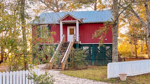 A red house on stilts with a blue roof, surrounded by trees and a white picket fence, with a path leading to the entrance.