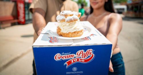 A person holds a box labeled "Cream Puffs To Go," featuring a large cream puff topped with whipped cream.