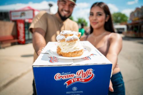 A man and woman hold a box of cream puffs, with a dessert topped with whipped cream in focus, against a sunny outdoor backdrop.