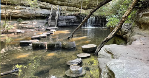 A serene natural scene featuring stepping stones in a calm waterway, surrounded by rocky cliffs and lush greenery.