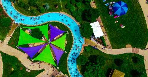 Aerial view of a winding lazy river with colorful shade sails and people enjoying the water and surrounding green areas.