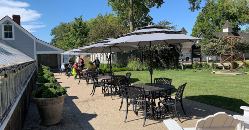 Outdoor seating area with tables and umbrellas, surrounded by greenery and people enjoying the space on a sunny day.