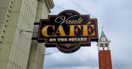 Sign for "The Vault Café on the Square" with a clock tower in the background under a cloudy sky.