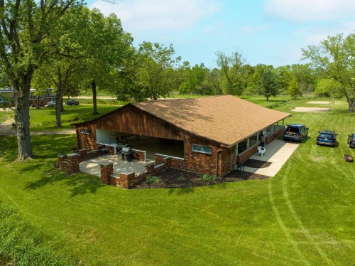 Aerial view of a brown-roofed building surrounded by green grass and trees, with parked cars nearby.