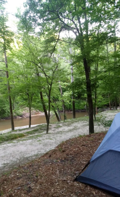 A serene campsite by a river, surrounded by lush green trees and sandy ground, with a tent in the foreground.