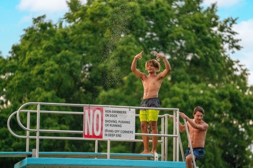 Two boys on a diving platform at a pool, one posing playfully while the other stands nearby. A "No" sign is visible.