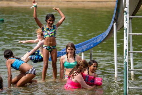 Children playing in a lake, some swimming and others on a slide, enjoying a sunny day outdoors.
