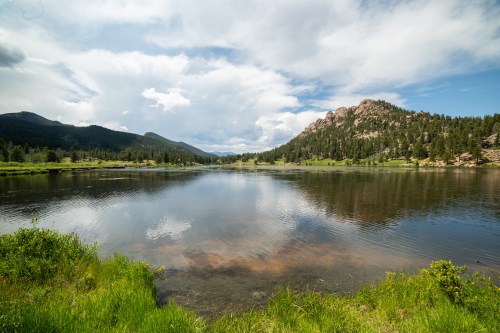 A serene lake surrounded by lush greenery and mountains under a partly cloudy sky. Reflections shimmer on the water.