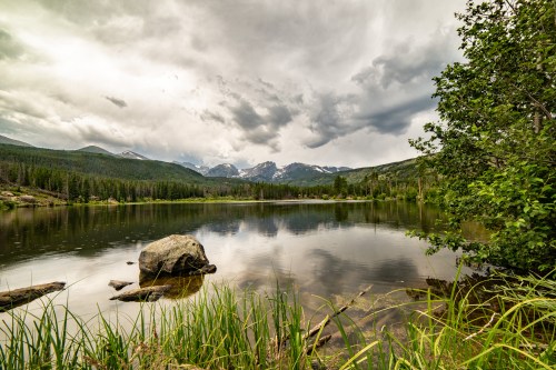 A serene lake surrounded by mountains and lush greenery under a cloudy sky. Reflections shimmer on the water's surface.