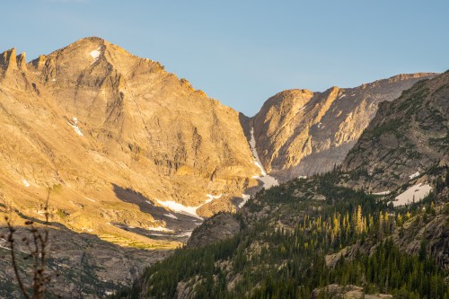 Majestic mountain landscape with rugged peaks, patches of snow, and lush green forests under a clear sky.