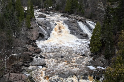 A rushing waterfall cascades over rocky terrain, surrounded by lush green trees and a rugged landscape.