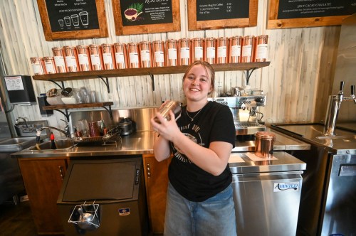 A smiling barista holds a shaker in a coffee shop, with cups displayed on the wall behind her.