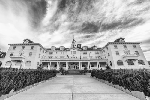 A grand, historic hotel with a symmetrical facade, surrounded by greenery, under a dramatic cloudy sky in black and white.