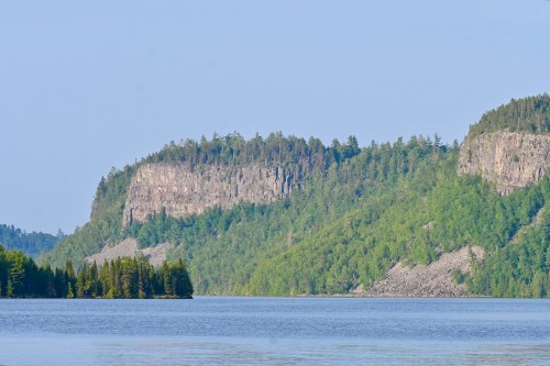A serene lake surrounded by lush green forests and rocky cliffs under a clear blue sky.