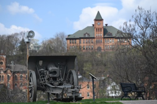 A vintage plow sits in the foreground with a historic building in the background under a partly cloudy sky.