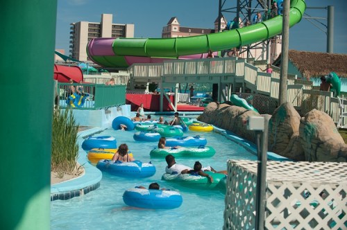 A water park scene with people floating on inner tubes in a lazy river, surrounded by slides and greenery.