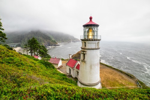 A lighthouse on a cliff overlooking a foggy ocean, surrounded by green hills and a cloudy sky.