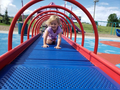 A young child crawls on a colorful playground structure, smiling under a bright blue sky.