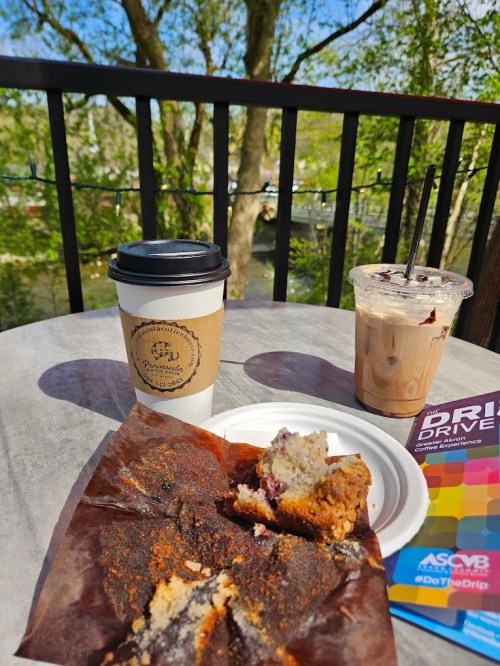 A coffee cup, iced drink, and a plate with a piece of muffin on a table outdoors, surrounded by greenery.