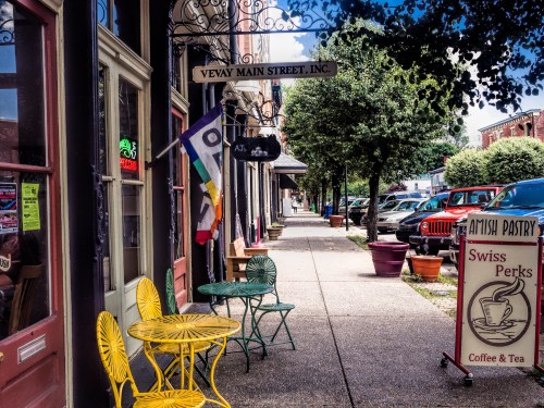 Colorful chairs line a quaint street with shops, featuring a rainbow flag and a sign for Swiss Perks Coffee & Tea.