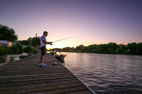 A person fishing from a wooden dock at sunset, with calm water and trees in the background.