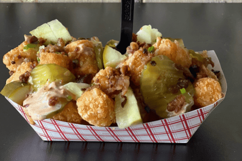 A tray of loaded tater tots topped with pickles, chopped meat, and a creamy sauce, served with a black fork.