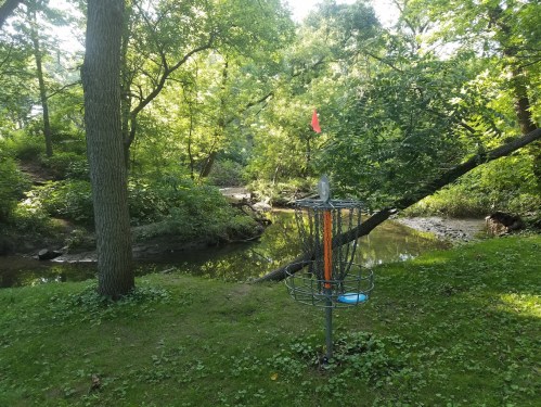 A disc golf basket stands near a serene creek, surrounded by lush greenery and trees.