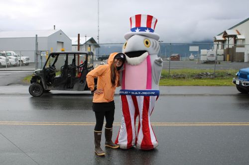 A person in an orange jacket poses with a large, cartoonish character dressed as Uncle Sam on a rainy street.
