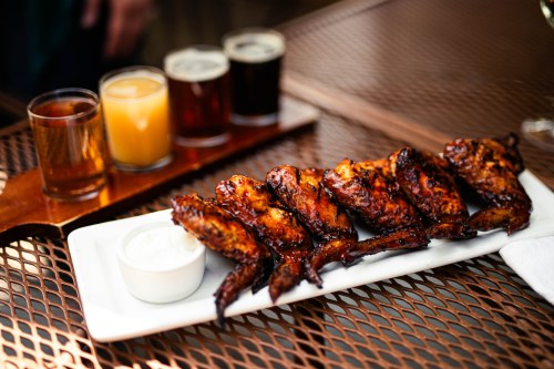 A platter of grilled chicken wings with a side of dipping sauce, accompanied by four small glasses of different beverages.