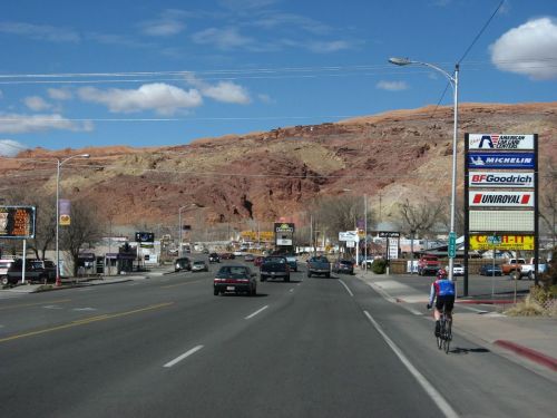 A cyclist rides along a road lined with shops, with red rock formations and blue skies in the background.