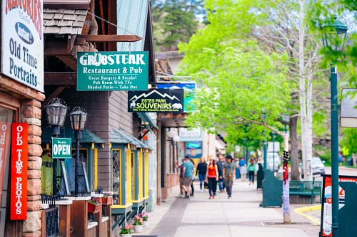 A lively street scene featuring shops and restaurants, with people walking and trees lining the sidewalk.