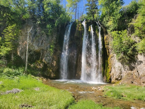 A serene waterfall cascading over rocky cliffs, surrounded by lush greenery and a clear stream in a sunny forest.