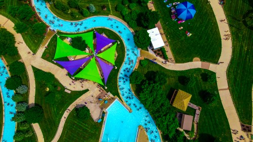 Aerial view of a water park featuring winding pools, colorful shade structures, and green lawns with visitors.