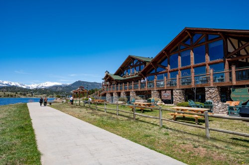 A scenic lakeside view featuring a rustic lodge, walking path, and mountains under a clear blue sky.