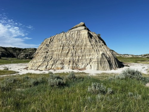 A large, layered rock formation rises from a grassy landscape under a clear blue sky.