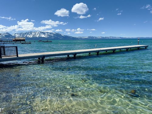 A wooden pier extends into clear blue water, with snow-capped mountains in the background under a bright blue sky.