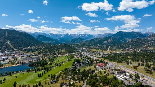 Aerial view of a scenic landscape featuring mountains, a lake, and a golf course under a blue sky with fluffy clouds.