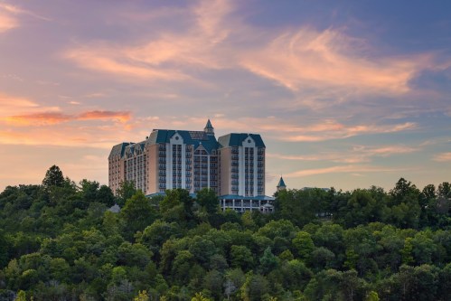 A large hotel building sits atop a hill, surrounded by lush green trees under a colorful sunset sky.