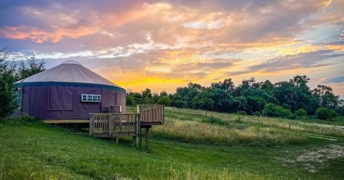 A yurt on a grassy hill at sunset, surrounded by trees and colorful clouds in the sky.