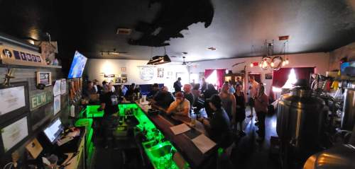 A lively bar scene with patrons seated at the counter and tables, featuring a green-lit bar and rustic decor.