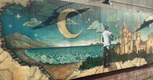 A vibrant mural depicting a coastal scene with mountains, a crescent moon, and a figure taking a photo.