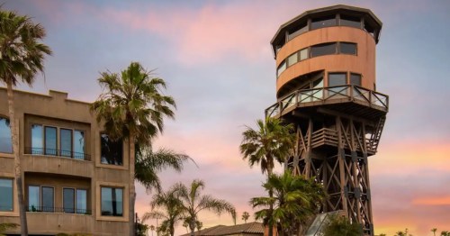 A modern observation tower beside a building, surrounded by palm trees against a colorful sunset sky.
