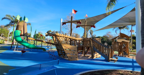 A colorful playground featuring a wooden pirate ship structure, slides, and a green caterpillar sculpture under a clear blue sky.