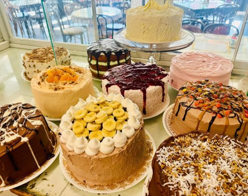 A display of various decorated cakes, including chocolate, fruit, and cream varieties, in a bakery setting.